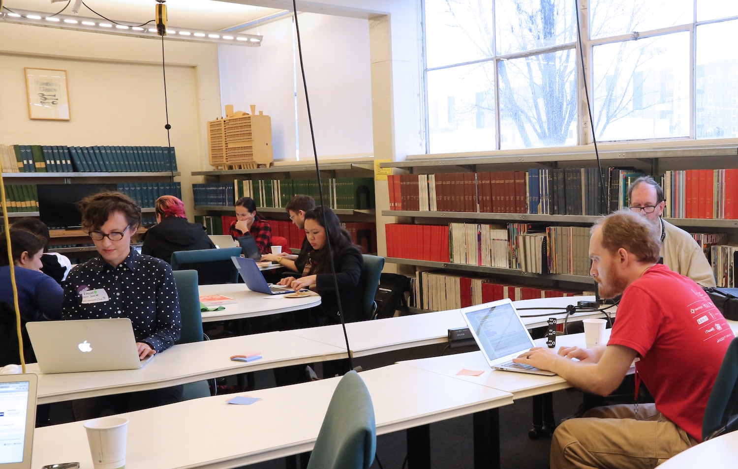 A group of people working on laptops in a library. Bound volumes with orange, green, and maroon spines are visible in the background. Two foreground figures are a dark-haired woman with a dotted shirt and a white man wearing a red Code4Lib shirt.